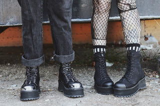 Two people wearing black Demonia platform boots, one with metallic toe caps and cuffed jeans, the other with matte finish boots, fishnet tights, and striped socks, standing on gritty urban ground.