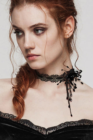 Woman wearing a black lace choker with a beaded side motif, styled with an off-shoulder black velvet dress against a plain background.
