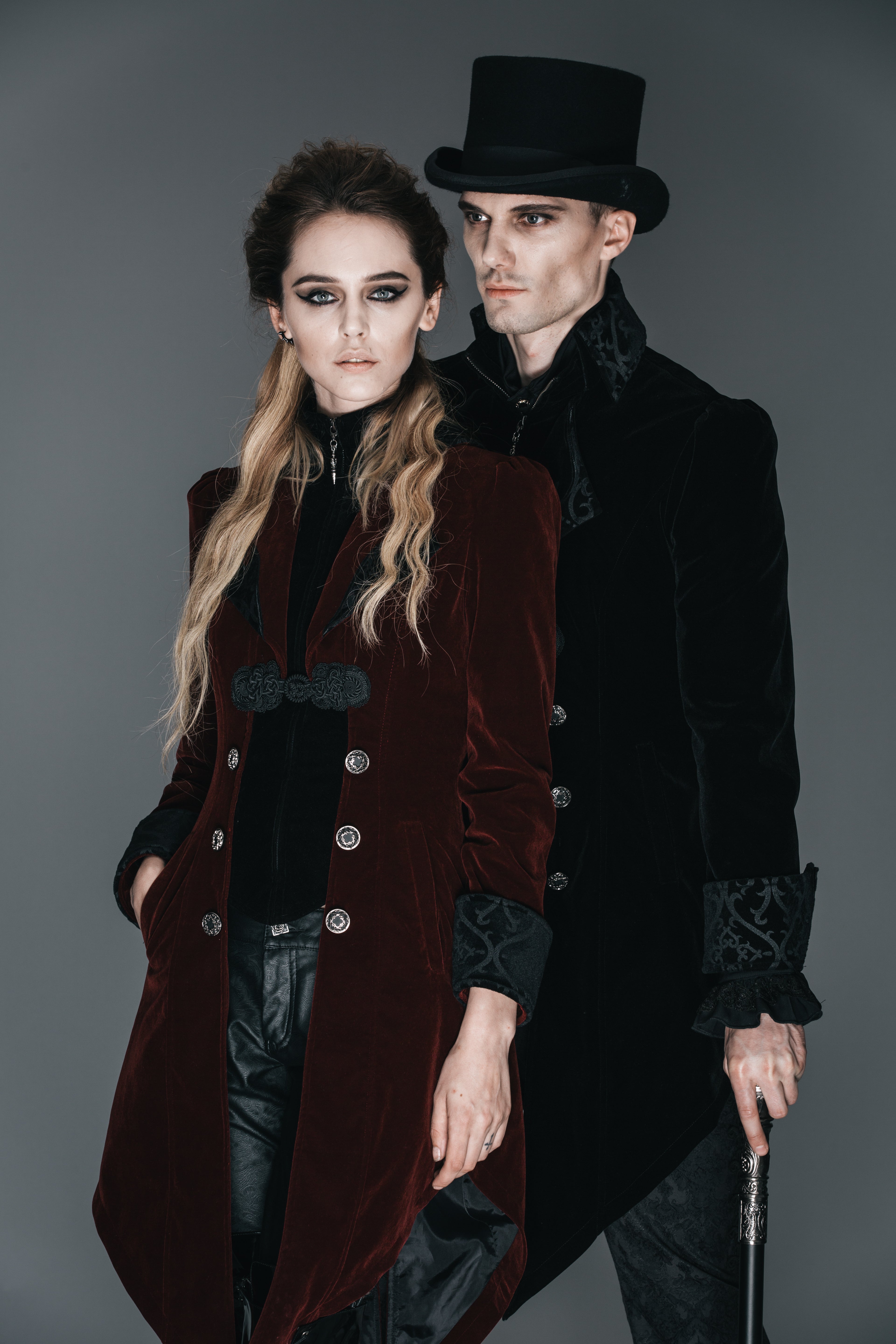 A Gothic wedding couple in Victorian-inspired black and burgundy outfits, posing against a dark background in elegant formalwear.