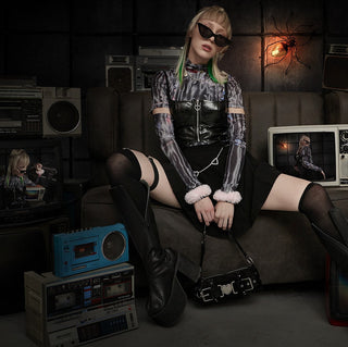 Young woman in platform boots and edgy ravewear sitting on a couch surrounded by vintage TVs and boomboxes, styled for a concert or festival night.