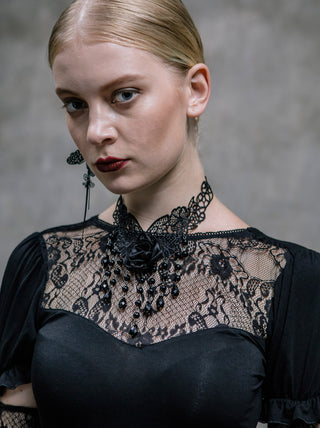 Woman wearing a black lace choker with a satin rose and beaded strands, styled with a sheer black lace top against a gray background.