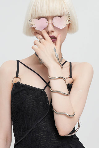 Woman wearing a silver snake-shaped accessory wrapped around her arm and neck, paired with a black strappy dress and pink heart-shaped sunglasses, on a white background.