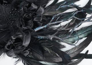 Close-up of black feathers, fabric roses, and branch details with blue accents on white background.
