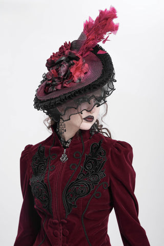 Side view of a woman in a red velvet Victorian jacket wearing a crimson feathered Gothic hat with lace veil and black rose trim, on a white background.