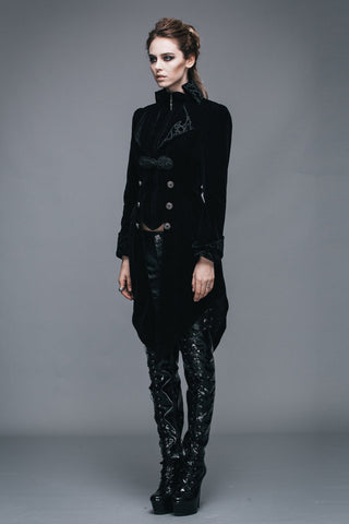 Full-body view of a woman wearing a long Gothic black velvet tailcoat with ornate buttons and asymmetrical hem, paired with shiny black lace-up platform boots and leather pants against a grey background.