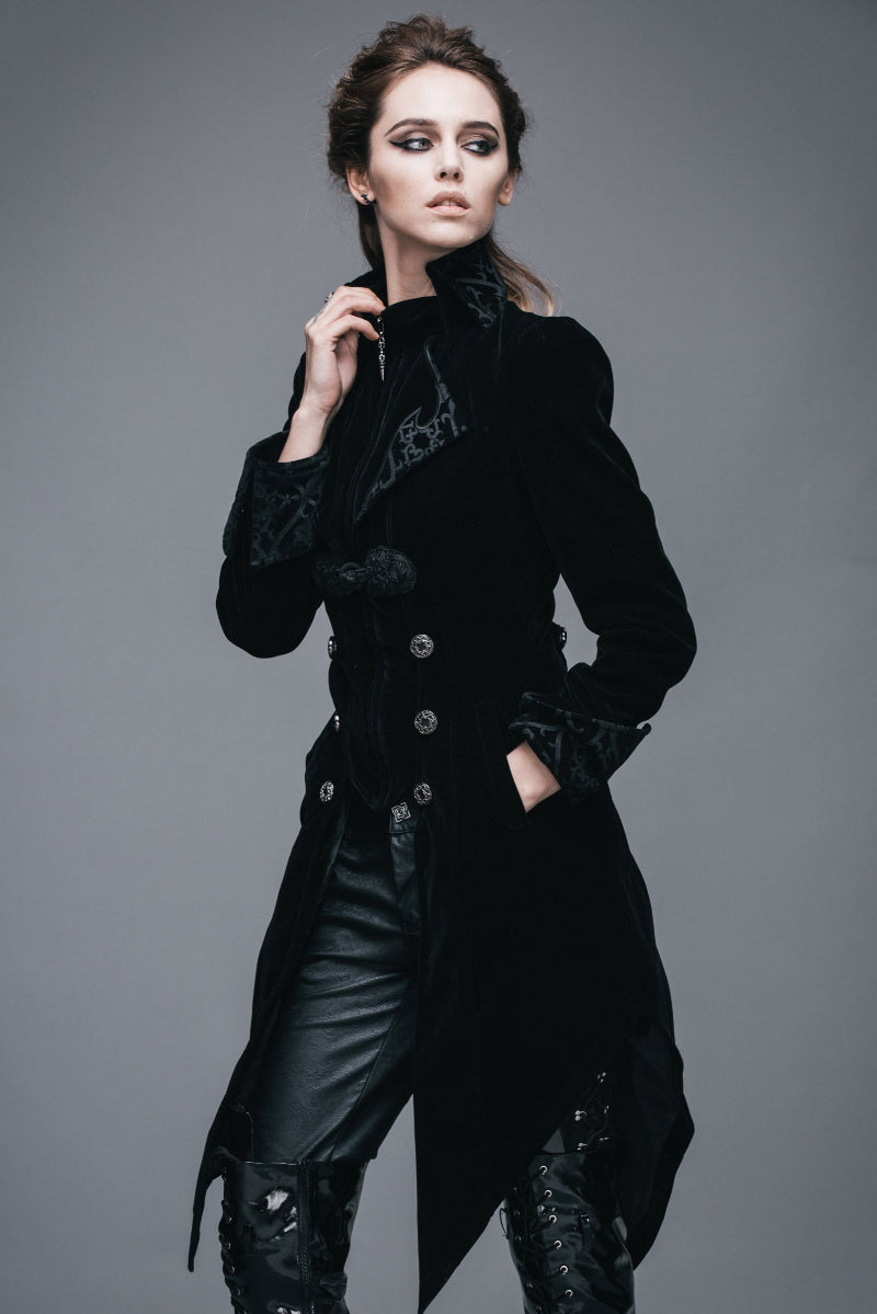 Woman in a high-collared Gothic black velvet tailcoat with decorative buttons, brocade lapels, and pointed hem, posing with one hand on her collar and the other in her pocket, wearing black lace-up pants.