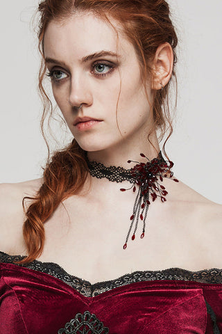 Woman wearing a red velvet off-shoulder dress with a black lace choker featuring a deep red 3D floral motif and cascading black beads.