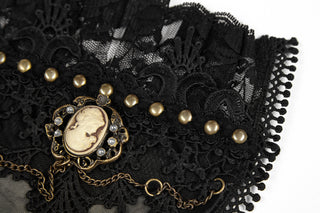 Close-up of black lace fabric with gold cameo brooch, pearl details, and gold bead trim, accented by a delicate gold chain, on a white background.